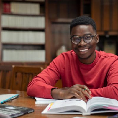 Portrait of university student doing homework in school library and smiling. Happy high school student looking at camera while studying for exam. African american clever guy with open book sitting at desk with copy space.